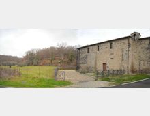 Edificio rurale in pietra con tetto a falde e piccole finestre, affacciato su un prato aperto. Un vialetto di ghiaia con cancello metallico conduce all’ingresso, con recinzione e alberi su una collina sullo sfondo.