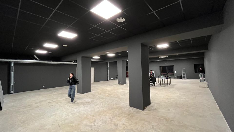 Ampio locale interno tipo magazzino o open space industriale con pareti grigio scuro, pavimento in cemento e colonne rettangolari. Soffitto a griglia nera con pannelli luminosi a LED e alcune attrezzature e tavoli sullo sfondo.