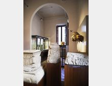 Sala museale con grandi sarcofagi e frammenti lapidei scolpiti esposti su piedistalli, affiancati da vetrine in vetro illuminate. L’ambiente ha pareti chiare, un’ampia arcata interna e una finestra alta sul fondo che lascia entrare luce naturale.