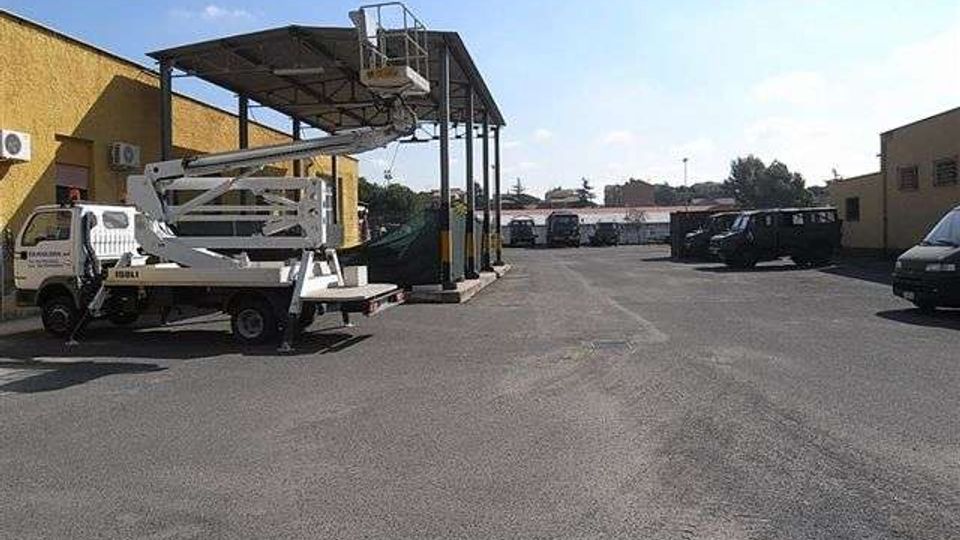 Ampio piazzale asfaltato in un’area industriale, con capannoni bassi ai lati e un’alta pensilina metallica al centro. Sono presenti veicoli da lavoro e parcheggiati lungo i bordi, tra cui un camion con piattaforma elevatrice.