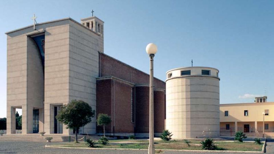 Chiesa moderna con facciata in pietra chiara e volumi geometrici, affiancata da una struttura cilindrica e da una torre campanaria con croce. Davanti si vede una piazza pavimentata con aiuole, piccoli alberi e un lampione, sotto un cielo sereno.
