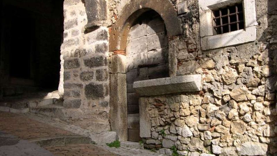 Facciata di un edificio in pietra con arco murato e portale ad arco, affiancato da una piccola finestra con inferriate. In primo piano si vede una strada lastricata in lieve salita e, a sinistra, un passaggio ad arco che conduce in una zona in ombra.
