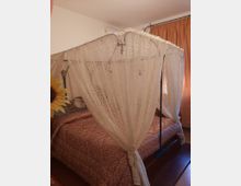 Camera da letto con letto matrimoniale coperto da un baldacchino in pizzo bianco fissato a una struttura metallica. Pareti chiare, pavimento in legno e finestra con tende arancioni sullo sfondo.