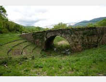 Ponte in pietra con un’unica arcata sopra un piccolo corso d’acqua, con parapetto metallico sulla sommità. Intorno si vede una valle erbosa con binari ferroviari dismessi curvi, alberi e colline sullo sfondo sotto un cielo nuvoloso.