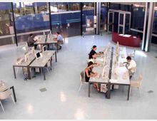 Ampio spazio interno tipo laboratorio informatico con pavimento chiaro, tavoli disposti in isole e numerosi computer desktop. Sullo sfondo ci sono pareti vetrate, porte d’ingresso e un bancone curvo vicino all’entrata.