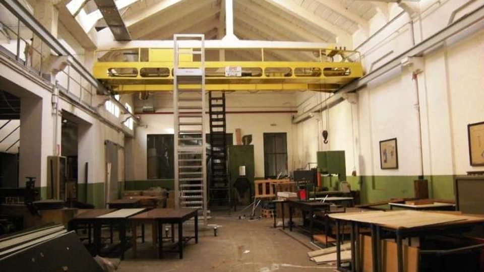 Interno di un capannone industriale adibito a officina, con soffitto alto a capriate e lucernari che illuminano lo spazio. Al centro è presente un carroponte giallo e una scala metallica, con banchi da lavoro e macchinari lungo le pareti.