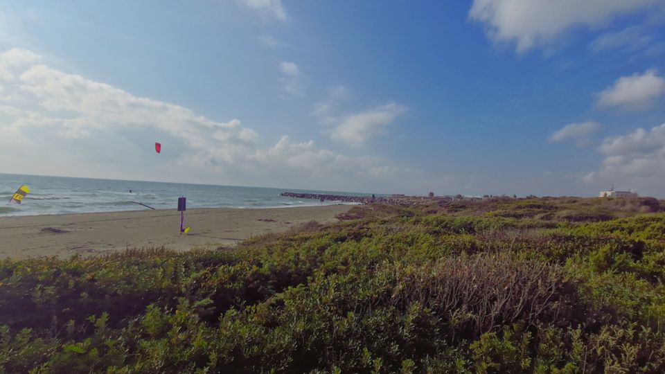 Veduta di una spiaggia sabbiosa affacciata sul mare con cielo parzialmente nuvoloso, delimitata in primo piano da dune basse ricoperte di vegetazione. In lontananza si nota un frangiflutti che si estende in acqua e alcuni piccoli edifici sulla costa.