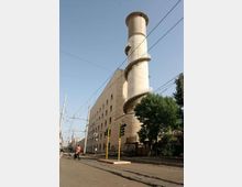 Edificio industriale in muratura chiara affacciato su una strada urbana con binari del tram e cavi aerei, dominato da un’alta torre cilindrica con scala a spirale esterna e ringhiere. In primo piano si vedono semafori e un albero lungo il marciapiede.