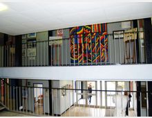 Interno di un edificio con atrio su due livelli, caratterizzato da passerelle con ringhiere metalliche e pareti espositive con quadri e cartelloni. Sul piano superiore è visibile un grande pannello artistico astratto e colorato, con finestre e porte vetrate sullo sfondo.