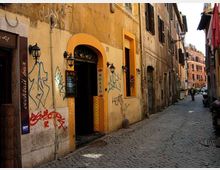 Vicolo urbano stretto con pavimentazione in sampietrini, fiancheggiato da edifici storici intonacati con finestre a persiane e lampioni a parete. In primo piano un ingresso ad arco con cornice arancione su una facciata con graffiti, mentre la strada prosegue tra palazzi fino a un incrocio sullo sfondo.