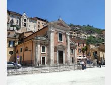 Facciata di una chiesa in mattoni e pietra con portale centrale e frontone triangolare sormontato da una croce, affacciata su una piazza con ringhiera. Sullo sfondo, case addossate a una collina e un campanile tra gli edifici.