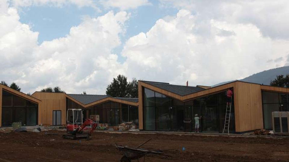 Complesso di edifici moderni in legno con tetti a falde e ampie vetrate, in un’area di cantiere con terreno sterrato e attrezzature da costruzione. Sullo sfondo si vedono alberi e colline sotto un cielo nuvoloso.