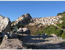 Veduta panoramica di un borgo arroccato su una collina rocciosa, con case in pietra addossate l’una all’altra e un edificio fortificato in cima. In primo piano si vedono un terreno ghiaioso con grandi massi e una panchina, circondati da vegetazione e cielo sereno.