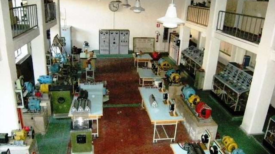 Interno di un laboratorio tecnico industriale con macchinari e motori esposti su banchi di lavoro, pavimento in mattoni rossi e pareti bianche alte. Lampade a sospensione, armadi elettrici e soppalchi con ringhiere corrono lungo i lati della sala.