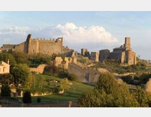 Panorama collinare con un castello medievale in pietra e mura merlate sulla sinistra e una chiesa in pietra con torre campanaria sulla destra. In primo piano si vedono bastioni e mura di cinta tra aree verdi, alberi e sentieri.