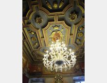 Soffitto a cassettoni decorato con cornici e rosoni dorati, con uno stemma centrale, in un ambiente interno di stile storico. Al centro è sospeso un grande lampadario in vetro con molte braccia illuminate, sopra pareti affrescate e finestre.