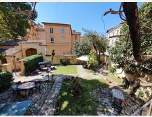 Cortile interno con area verde e pavimentazione in pietra, delimitato da muri e circondato da edifici in intonaco chiaro. Sono presenti alberi e siepi, tavoli e sedie da esterno, vasi con piante e un gazebo/ombrellone su una zona lastricata.
