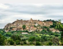 Borgo collinare con case in pietra addossate tra loro e una chiesa con campanile in cima, circondato da vegetazione. In primo piano si vede una valle verde con alberi e campi sotto un cielo nuvoloso.