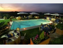 Piscina rettangolare all’aperto illuminata al crepuscolo, circondata da prato e da tavoli apparecchiati con tovaglie e sedie. Attorno ci sono ombrelloni bianchi, fioriere e muretti in pietra, con vista su una campagna collinare sullo sfondo.