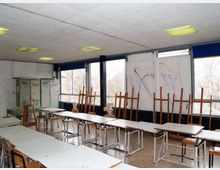 Aula scolastica vuota con file di banchi e sedie, armadietti metallici e pareti con segni e graffiti. Lungo un lato ci sono grandi finestre con infissi scuri e pannelli oscuranti blu, con cavalletti da disegno appoggiati vicino ai vetri.