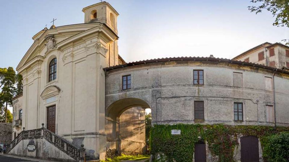 Facciata di una chiesa storica intonacata chiara con campanile e scala d’accesso, affiancata da un edificio adiacente curvilineo a due piani con finestre e portico ad arco. In primo piano si vedono muri parzialmente coperti d’edera e un piccolo passaggio sotto l’arco in un contesto urbano con alberi ai lati.