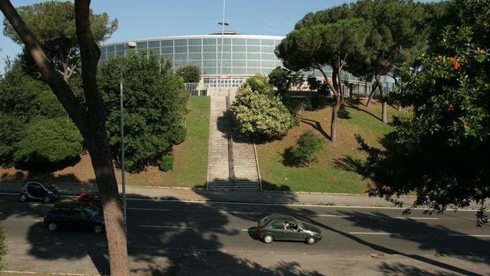 Edificio moderno a pianta semicircolare con ampia facciata vetrata, situato in cima a una collina. Una lunga scalinata centrale attraversa un prato alberato e conduce all’ingresso, con una strada trafficata in primo piano.