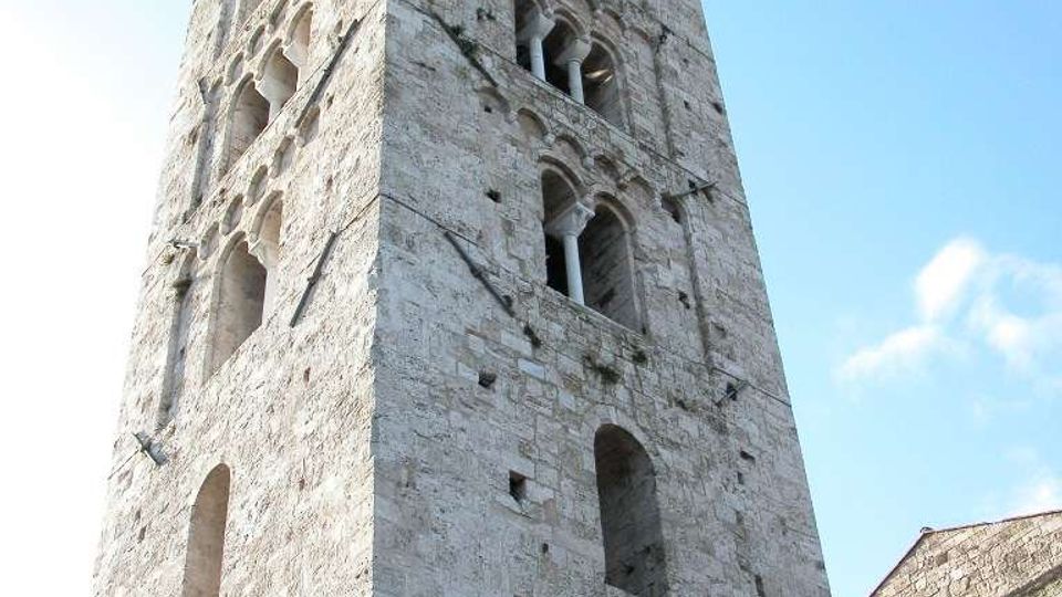Torre campanaria medievale in pietra chiara vista dal basso, con più ordini di bifore e trifore ad arco e muratura massiccia. Sul lato destro si intravede parte di un edificio in pietra con finestre ad arco sotto un cielo azzurro.