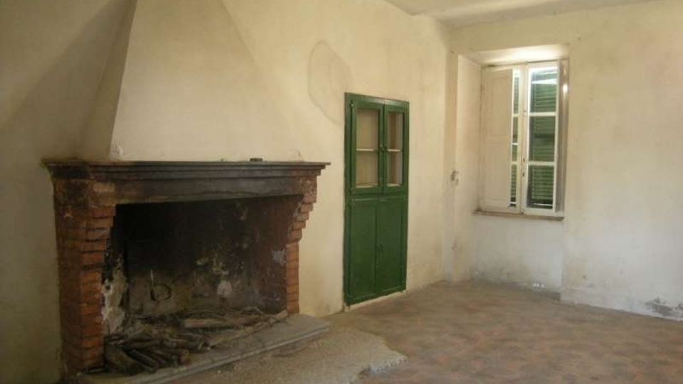Stanza interna spoglia con pareti intonacate chiare e pavimento in cotto, con un grande camino in muratura e mattoni sul lato sinistro. Sul fondo si vedono una porta verde e una finestra alta con persiane verdi.