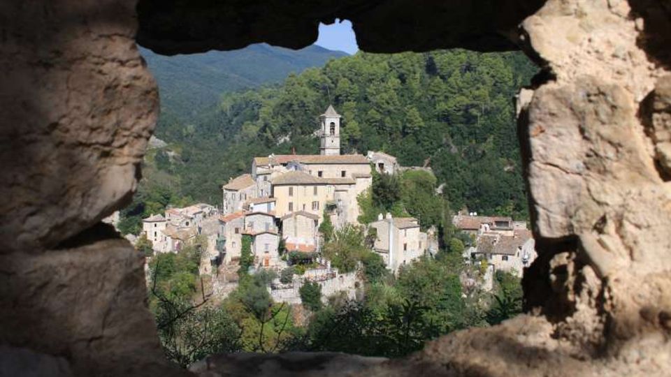 Veduta di un borgo collinare con case in pietra e un campanile centrale, incastonato tra boschi e montagne. Il paesaggio è inquadrato da un’apertura irregolare in un muro di pietra in primo piano.