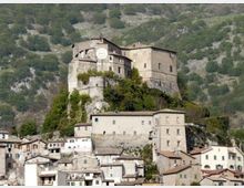 Castello in pietra su uno sperone roccioso sopra un borgo, con un edificio principale dotato di grande orologio sulla facciata e mura di contenimento. Sullo sfondo si vede un pendio montano con vegetazione rada.