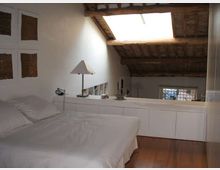 Camera da letto in soppalco con letto matrimoniale, armadiature bianche basse e lampada da tavolo. Soffitto mansardato con travi in legno e grande lucernario, pavimento in legno e finestra sulla parete di fondo.