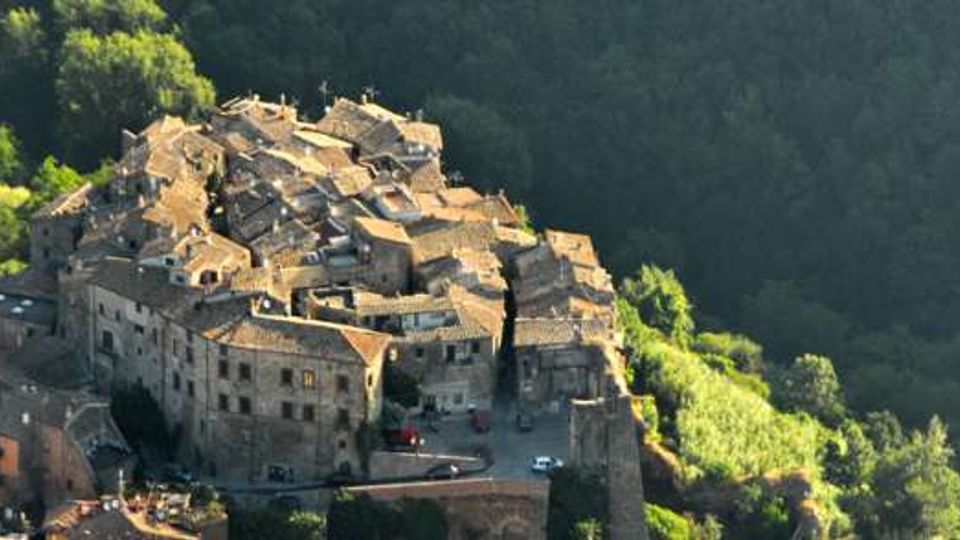 Veduta aerea di un borgo medievale arroccato su una collina, con case in pietra addossate tra loro e tetti in tegole. Il paese è circondato da un fitto bosco verde e si nota una cinta muraria lungo il margine dell’abitato.