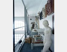 Corridoio in un edificio adibito a deposito o laboratorio museale, con passerella metallica affacciata su grandi finestre ad arco. Lungo la parete sono disposte statue e bassorilievi, con quadri appoggiati su tavoli e alcune scatole sul pavimento.