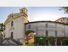 Facciata di una chiesa storica intonacata chiara con campanile e scala d’accesso, affiancata da un edificio adiacente curvilineo a due piani con finestre e portico ad arco. In primo piano si vedono muri parzialmente coperti d’edera e un piccolo passaggio sotto l’arco in un contesto urbano con alberi ai lati.