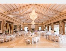 Ampia sala per eventi con soffitto alto a capriate in legno bianco e grandi lampadari a goccia centrali. Tavoli rotondi apparecchiati con sedie bianche, ampie finestre con tende laterali e camino sul fondo della sala.