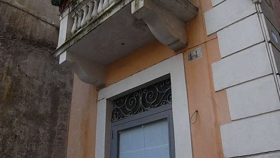 Facciata di un edificio urbano con balcone in muratura e balaustra, decorato con fioriere e vegetazione. Sotto il balcone si vede un portone alto con inferriata ornamentale, pareti intonacate e una targa rotonda sul muro laterale.