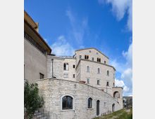 Edificio storico in pietra chiara, simile a un castello o monastero, con più livelli e finestre ad arco, visto dal basso dietro un alto muro in pietra. Sullo sfondo si vede il cielo azzurro con nuvole e un sentiero erboso lungo il perimetro.