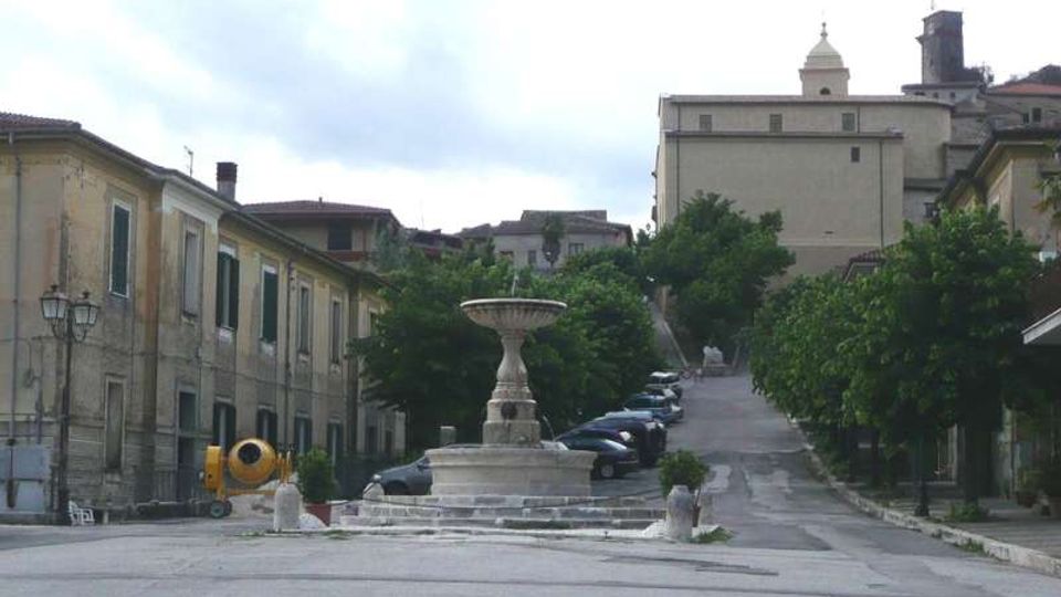 Piazza di un borgo con fontana in pietra al centro e strada in salita che prosegue tra alberi e auto parcheggiate. Ai lati si vedono edifici storici a due piani con persiane e lampioni, con un grande edificio in cima alla salita.