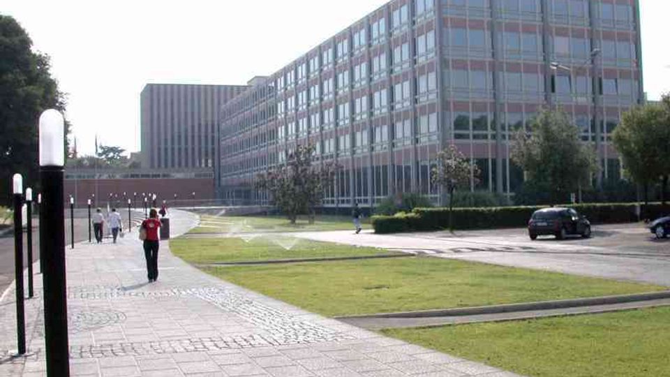 Viale pedonale pavimentato che attraversa un’area verde in un complesso di uffici, con grandi edifici moderni a più piani e facciate a griglia di finestre. A lato si vedono lampioni, siepi e una strada interna con alcune auto parcheggiate.
