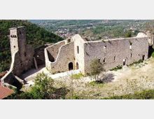 Rovine di un castello in pietra su un pendio, con una torre quadrata, alte mura con piccole finestre e un cortile interno in parte aperto. Sullo sfondo si vede una valle verde con un centro abitato e colline circostanti.