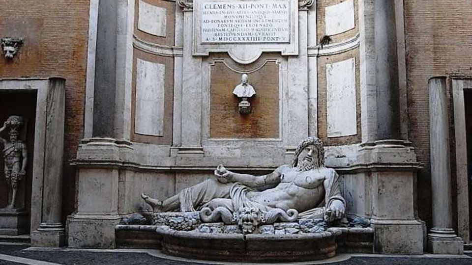 Fontana monumentale in stile barocco incassata in una parete di mattoni, con colonne laterali, timpano curvilineo e grande iscrizione centrale. In basso una vasca semicircolare con statua reclinata in marmo, su una piazzetta pavimentata a raggiera.