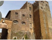 Edificio storico in muratura dall’aspetto di torre fortificata, con facciata in intonaco consumato e finestre piccole ad arco. Sulla destra si vede una torre quadrangolare in mattoni con merlature in cima, collegata a un arco d’ingresso laterale.