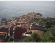 Vista panoramica di un borgo collinare con case addossate e tetti in coppi rossi, dominato da un grande edificio storico su un’altura. Sullo sfondo si estende una valle con campi e vegetazione, in lontananza leggermente velata.