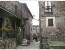 Vicolo acciottolato in un borgo storico, stretto tra edifici in pietra e intonaco con balconi in ferro battuto, persiane in legno e un lampione a muro. In fondo si vede un passaggio ad arco e una targa stradale su una facciata.