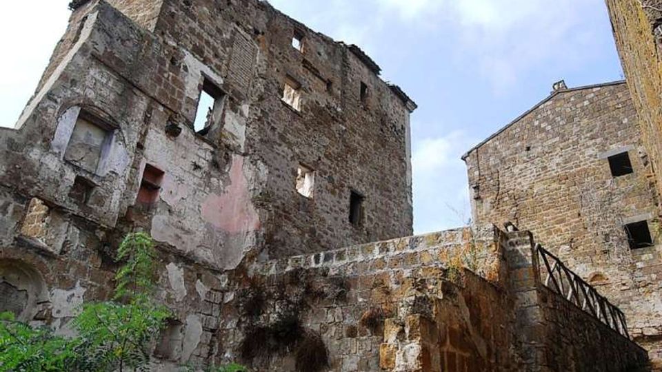 Ruderi di un edificio in pietra a più piani, con muri scrostati e finestre senza infissi, affacciati su un cortile con vegetazione. In primo piano si vede un muro di contenimento in pietra e una scala esterna con ringhiera che sale verso un altro fabbricato in muratura.