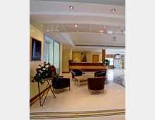 Hall di un hotel con pavimento in piastrelle lucide e area salotto con quattro poltrone scure e tavolino basso al centro. Sullo sfondo si vede il banco reception in legno, illuminazione a faretti nel controsoffitto, vetrate laterali e decorazioni come un grande vaso di fiori e quadri alle pareti.