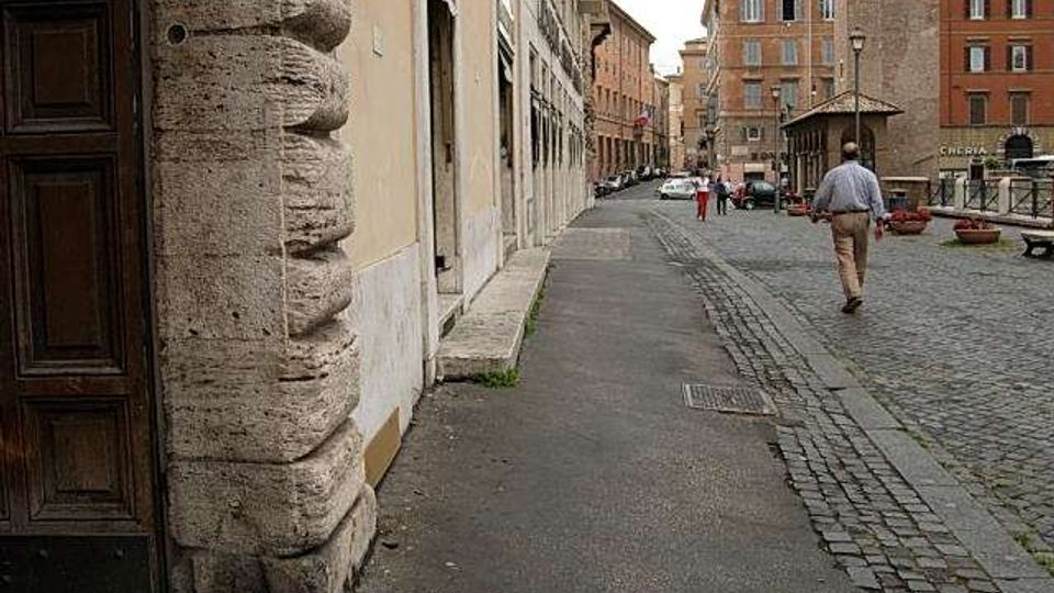 Strada urbana acciottolata con marciapiede stretto, fiancheggiata da edifici in muratura e intonaco con portoni e finestre. Sullo sfondo si vedono palazzi di mattoni, un’alta struttura in pietra tipo torre e una piccola area con panchine e fioriere.