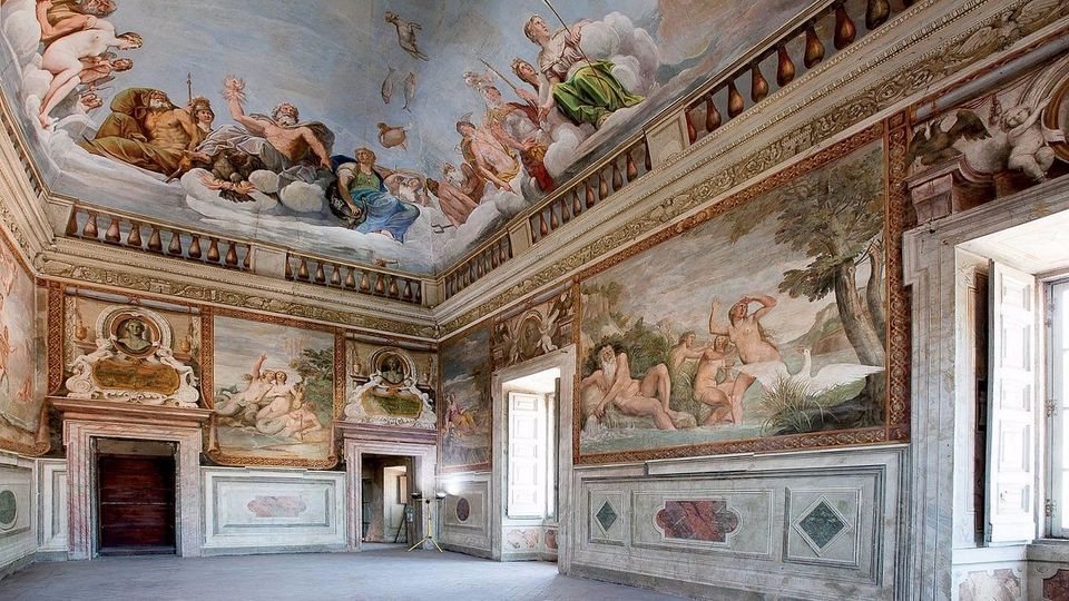 Ampia sala storica con pareti e soffitto interamente affrescati con scene mitologiche e figure tra nuvole, bordure decorative e pannellature in stile classico. L’ambiente ha pavimento in cotto, grandi finestre con scuri bianchi e porte incorniciate da stucchi e dipinti.