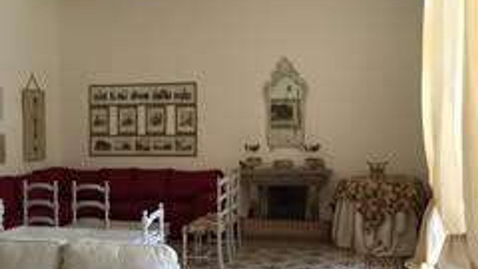 Salotto in stile tradizionale con divano chiaro, pareti bianche con decori e cornici, e pavimento a piastrelle con motivo geometrico e bordura. Ampie tende chiare filtrano la luce su arredi d’epoca, tra cui sedie in legno, un camino e un tavolino.