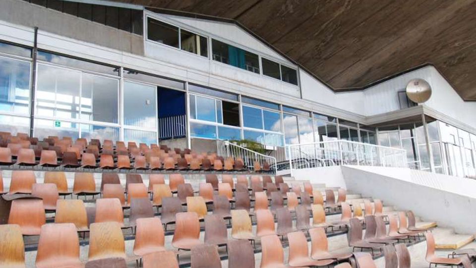 Gradinate di uno stadio o arena con file di sedili in plastica marrone disposti su più livelli. Sullo sfondo si vede una struttura coperta con grandi vetrate, ringhiere metalliche e un soffitto inclinato in cemento.